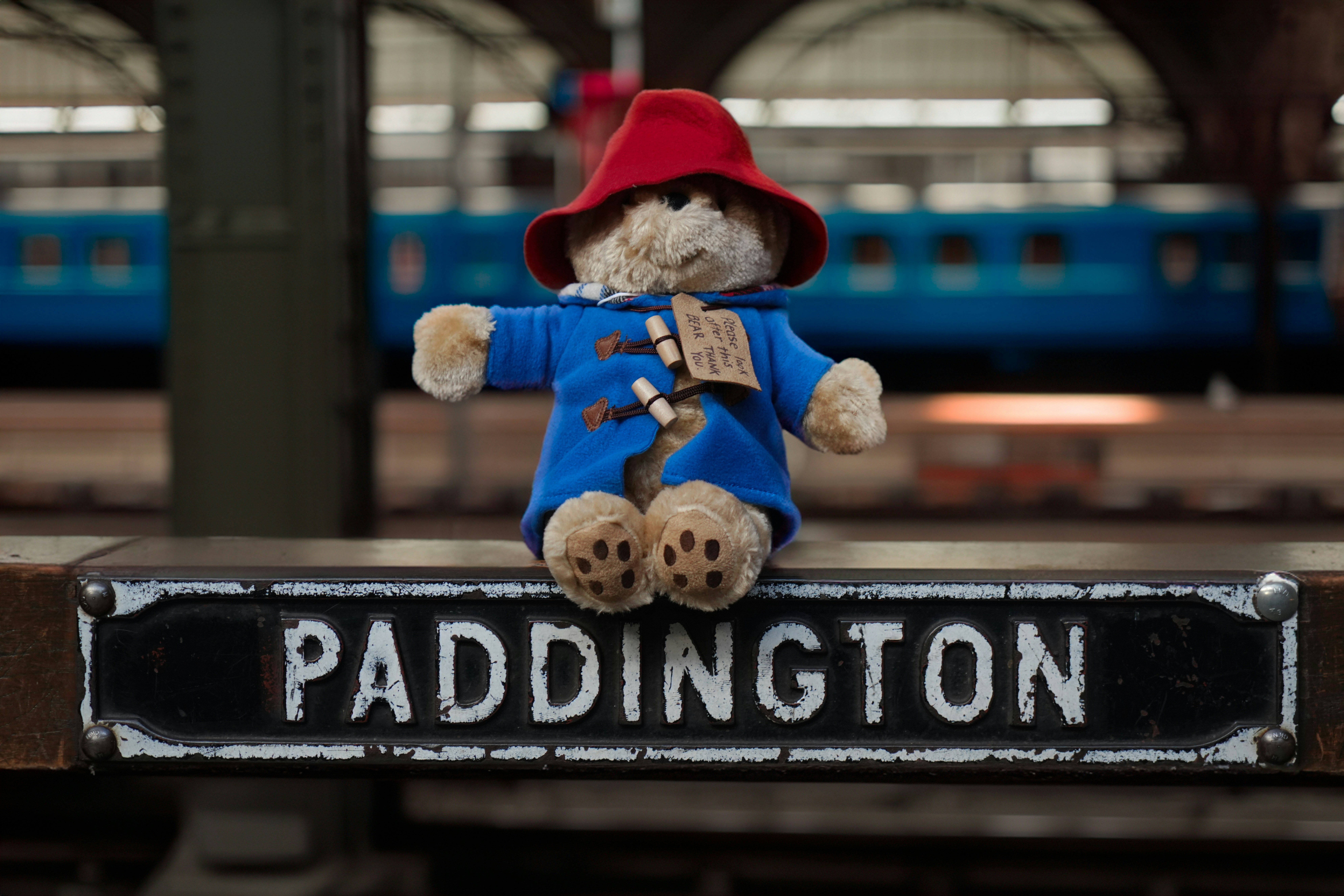 Paddington teddy bear gift for the Christmas audiobook gift guide A photo of a Paddington teddy bear placed on top of a sign which reads Paddington, inside Paddington station in London