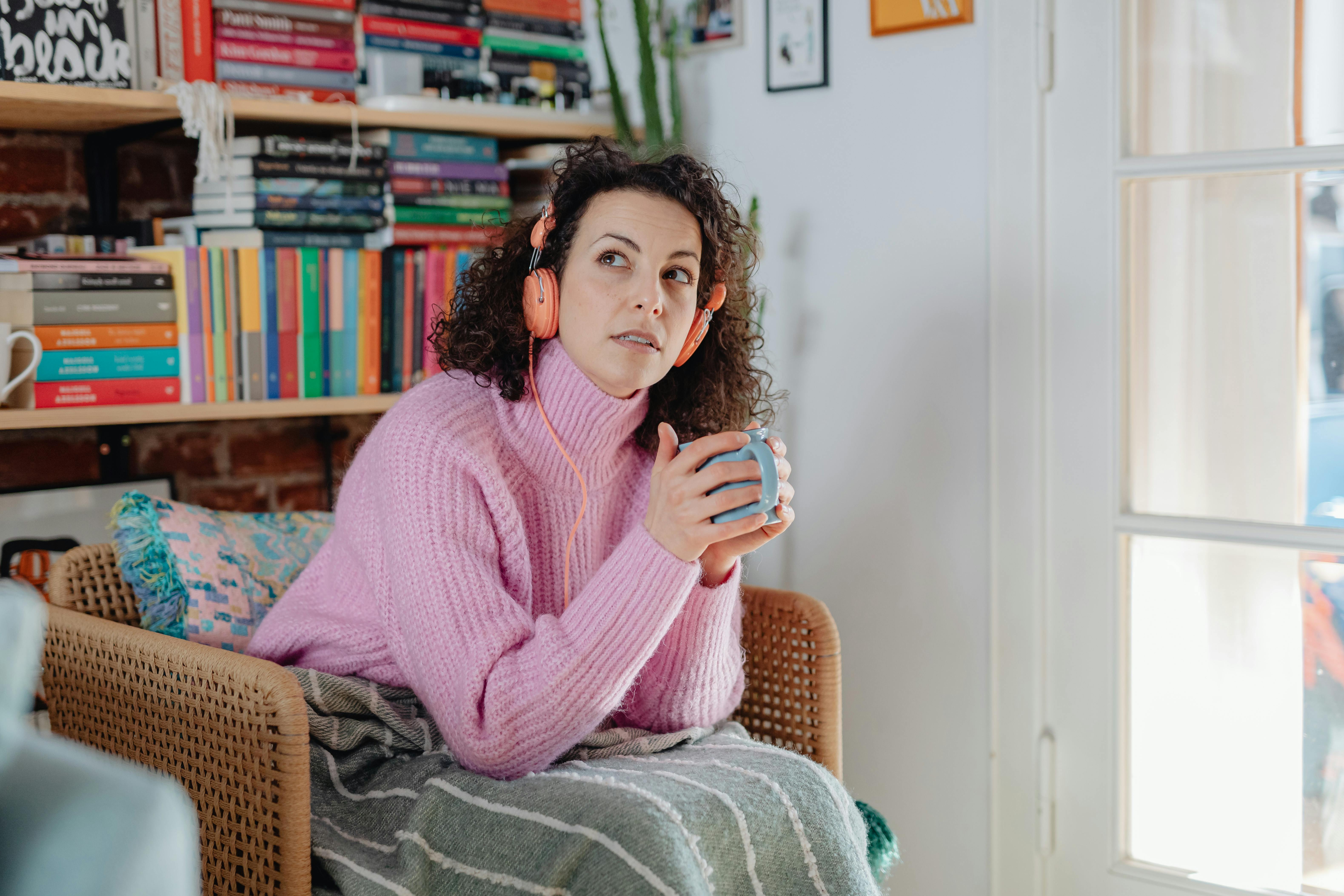 Grey and white striped throw A woman sitting in a chair, holding a mug with pink headphones on. She has a grey blanket with white stripes over her leg. She is sitting in a living room with a bookcase behind her.