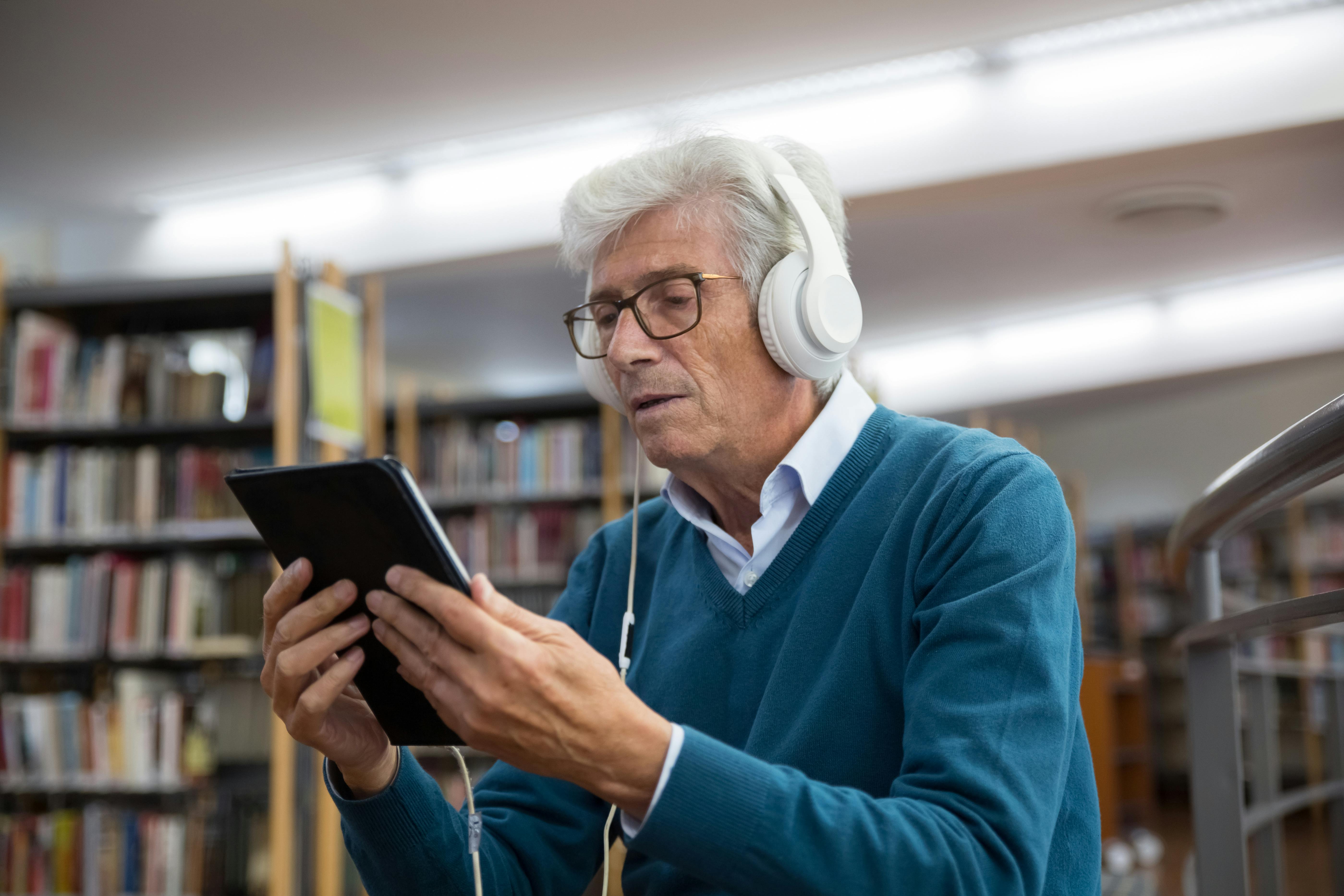 A photo of an older gentleman with headphones on holding a tablet in a library