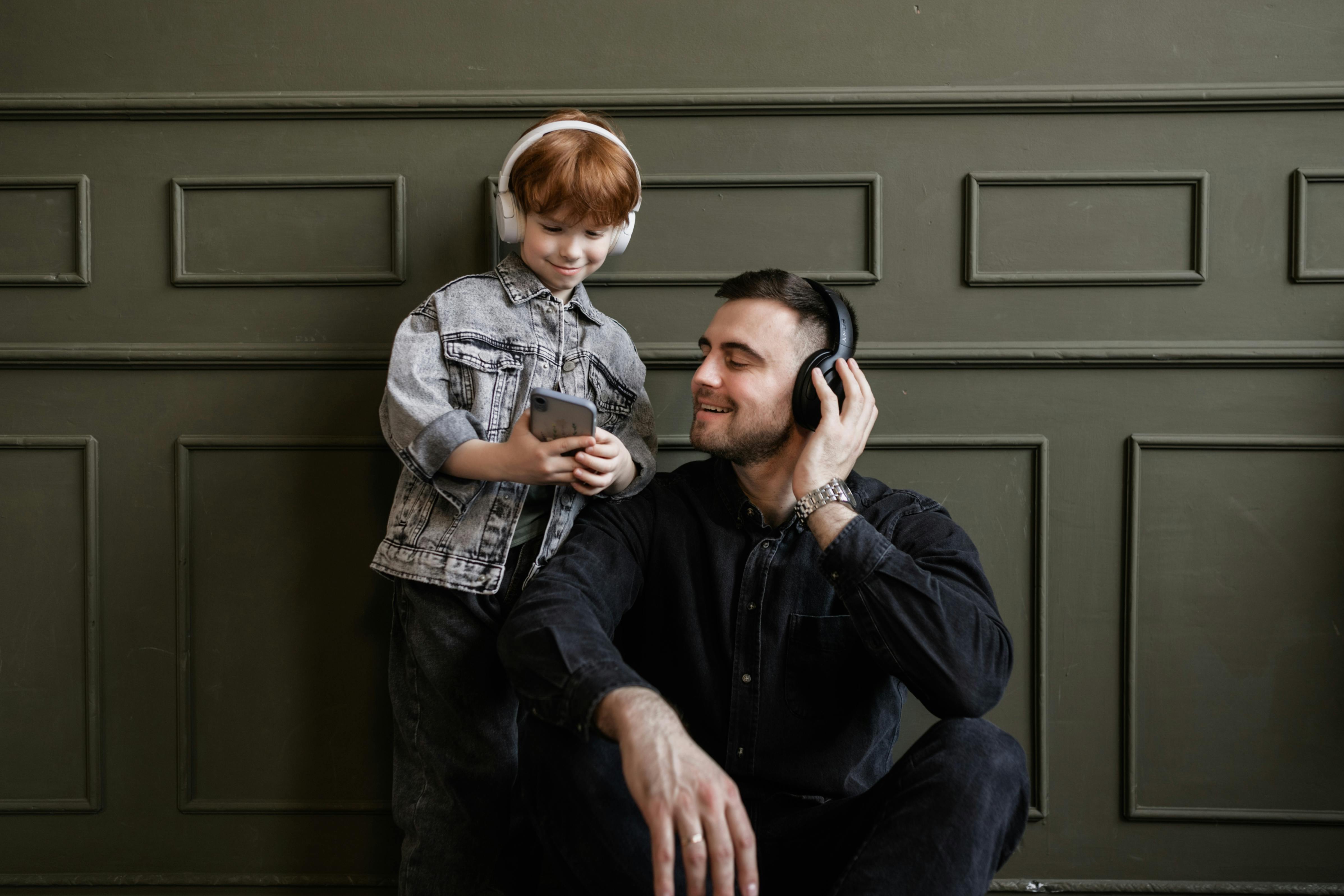A photo of a father and son and both have headphones on while looking at the phone the son is holding 