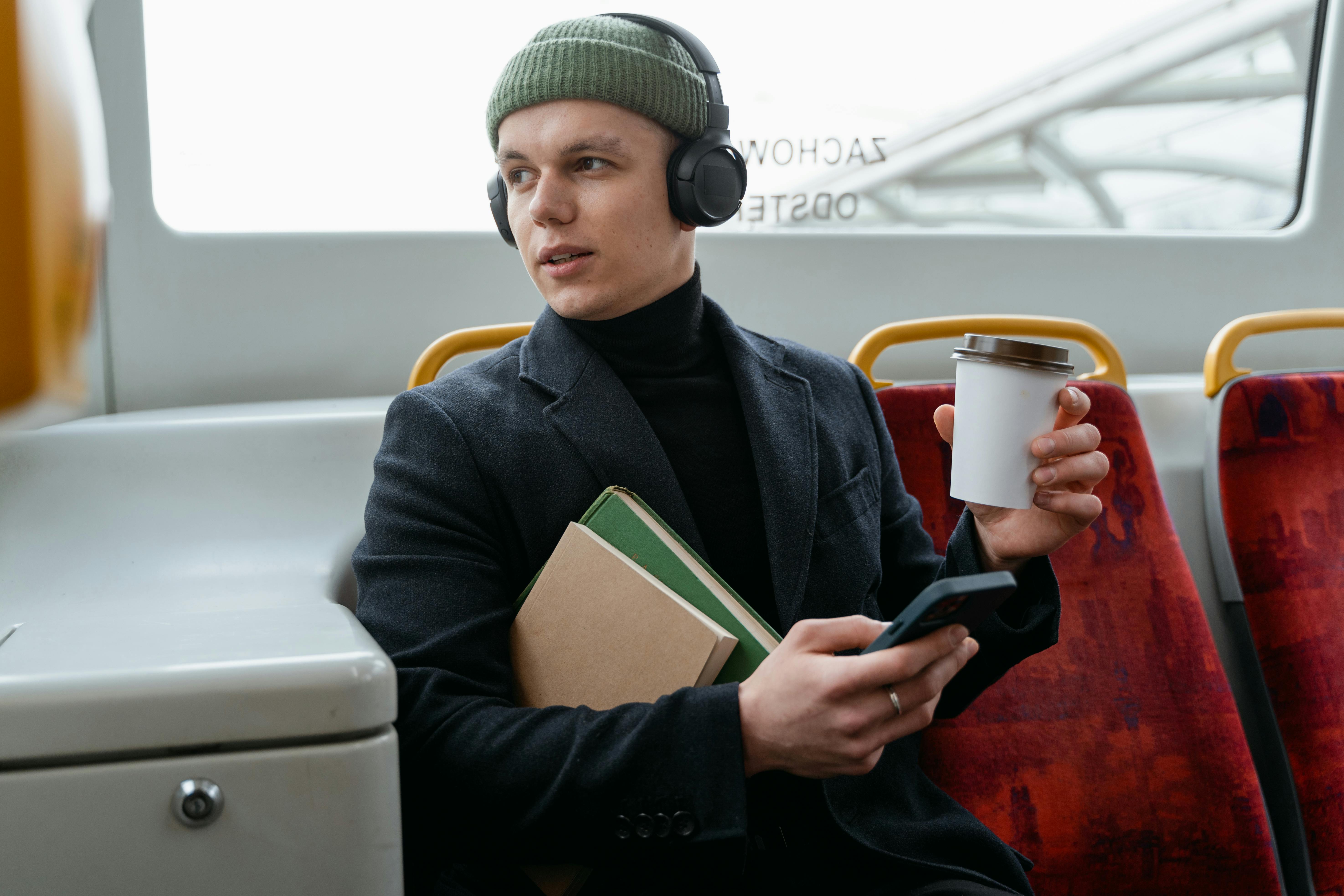 A man wearing a hat, holding books and a coffee while listening to headphones