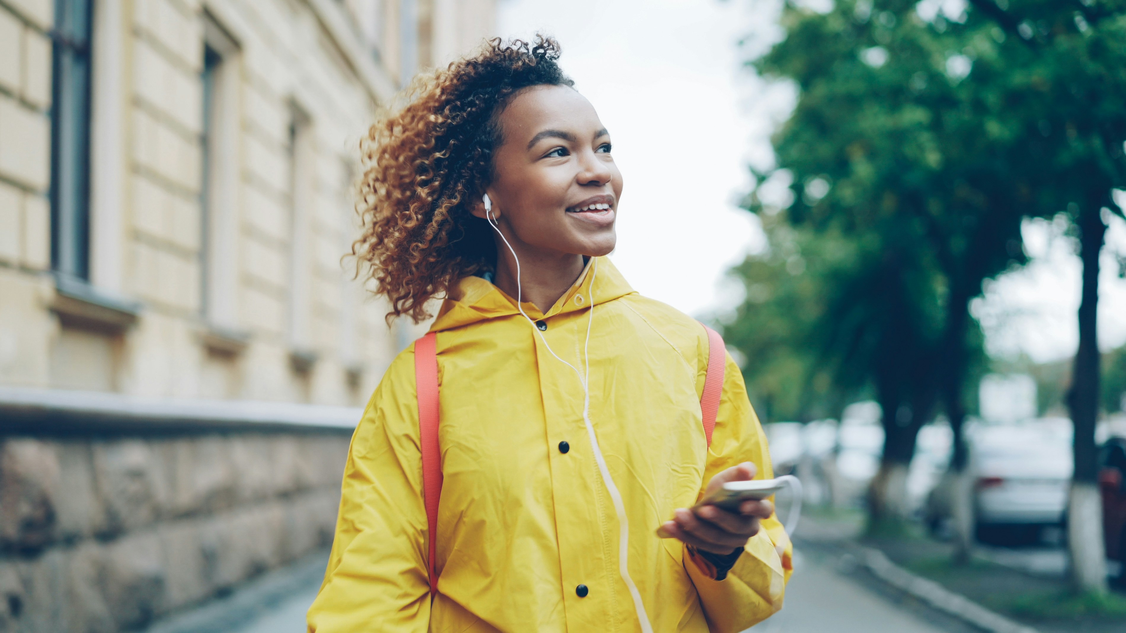 A woman wearing a yellow raincoat walking down a road holding a phone and wearing headphones