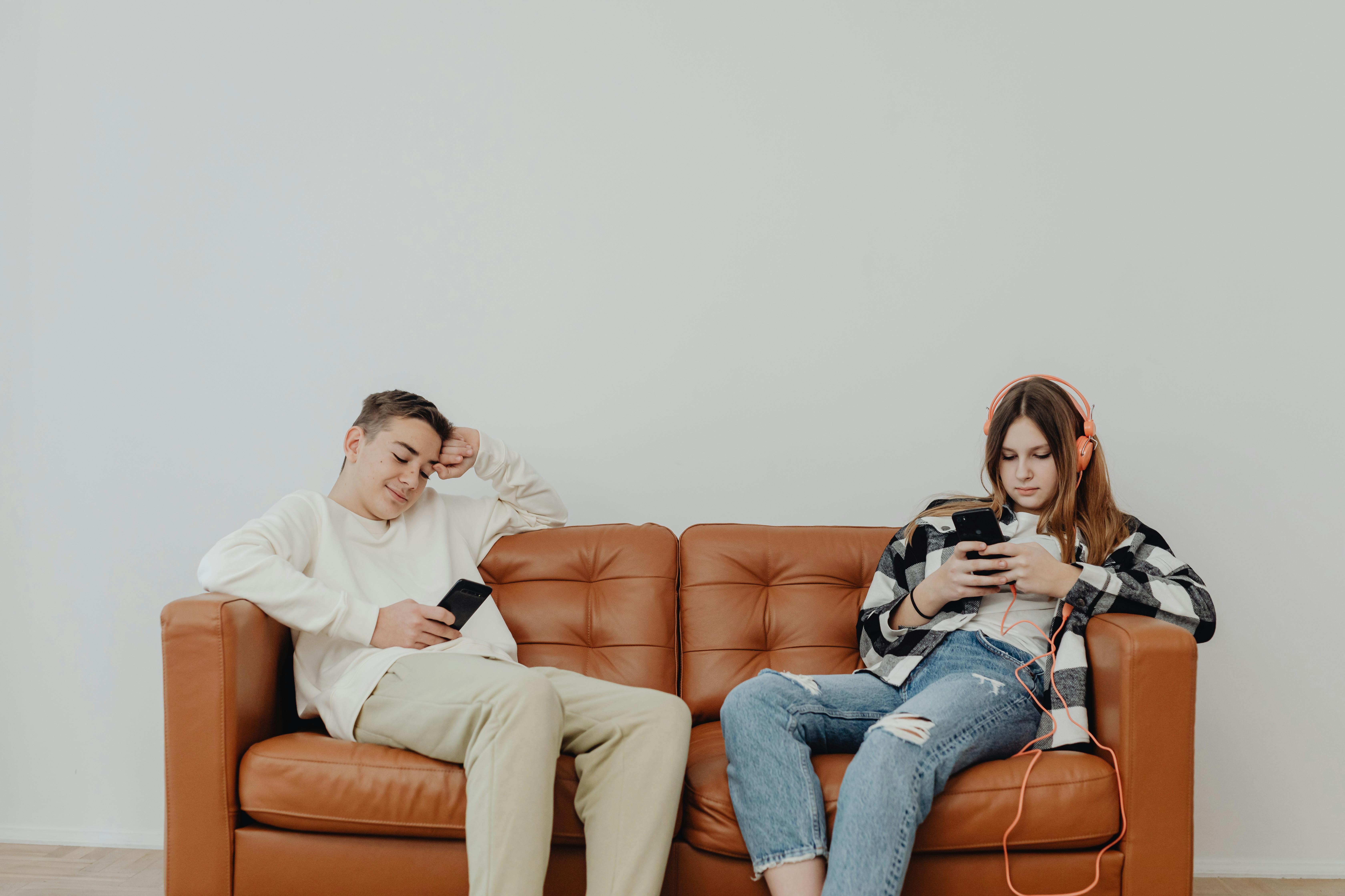A photo of two teens sitting on an orange sofa looking at their phones with headphones on