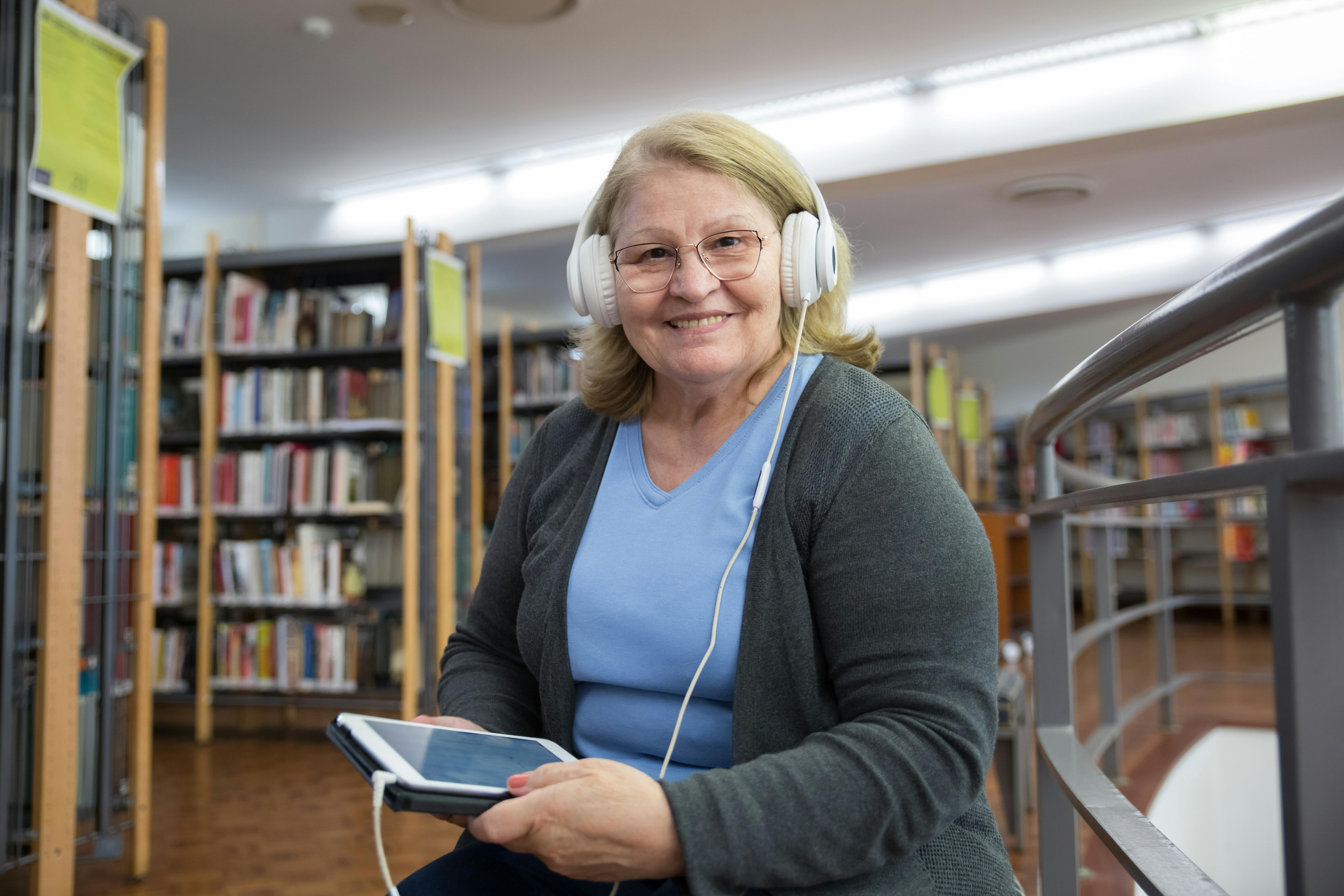 A photo of an older woman in a library with headphones on holding a tablet